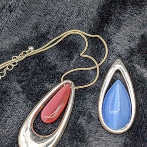 May include: Two silver-toned necklaces with teardrop pendants. One pendant features a red oval stone within a larger teardrop frame. The other pendant has a blue teardrop stone. The necklaces are on a dark, textured surface.