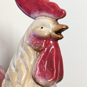 May include: A ceramic rooster figurine with a red comb and wattle. The rooster's body is cream-colored with brown accents, and the beak is brown. The figurine is glossy and appears to be hand-painted.