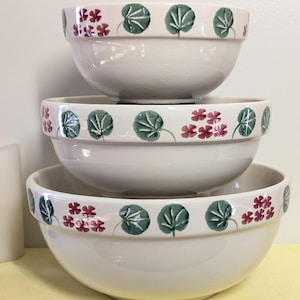 May include: A stack of three off-white ceramic mixing bowls. Each bowl has a decorative band around the rim featuring green leaves and small red flowers. The bowls are nested, showcasing their varying sizes and the floral design.
