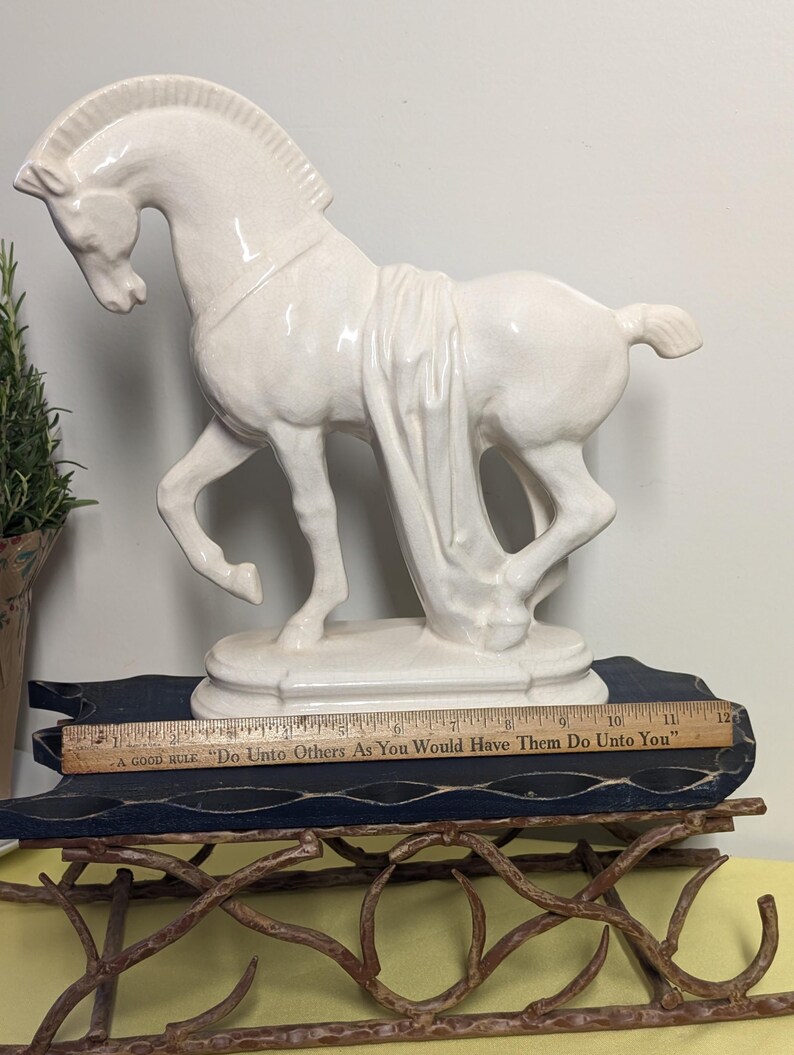 Puede incluir: Una estatua de caballo de cer&aacute;mica color crema con detalles de tela drapeada. El caballo est&aacute; posado con una pata levantada, montado sobre una base redonda. Una regla con el texto "A GOOD RULE 'Do Unto Others As You Would Have Them Do Unto You'" es visible.