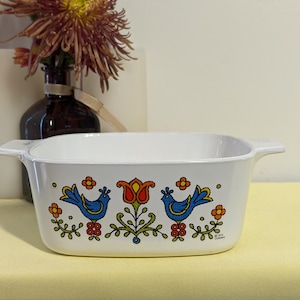May include: White rectangular casserole dish with handles, featuring a colorful design of blue birds, orange flowers, and a red tulip. The dish has a vintage aesthetic, perfect for serving food.