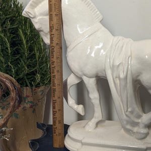 Puede incluir: Una figura de caballo de cer&aacute;mica blanca, de aproximadamente 28 cm de altura, se encuentra sobre una base decorativa. El caballo est&aacute; en movimiento, con detalles precisos. Una regla de madera se coloca junto al caballo. El texto en la regla dice: "A GOOD RULE 'Do Unto Others As You Would Have Them Do Unto You'".