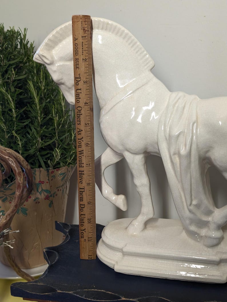 Puede incluir: Una figura de caballo de cer&aacute;mica blanca, de aproximadamente 28 cm de altura, se encuentra sobre una base decorativa. El caballo est&aacute; en movimiento, con detalles precisos. Una regla de madera se coloca junto al caballo. El texto en la regla dice: "A GOOD RULE 'Do Unto Others As You Would Have Them Do Unto You'".