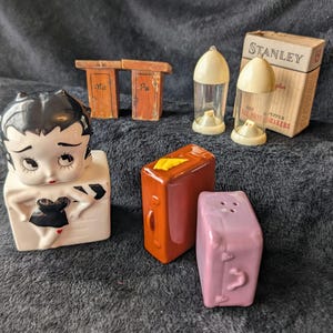 May include: A set of vintage salt and pepper shakers featuring a cartoon character, a suitcase, and a pair of outhouses. The shakers are made of ceramic and have a painted finish. The set includes a box labeled "Stanley Plus Salt & Pepper Shakers."