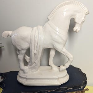 Puede incluir: Figura de caballo de cer&aacute;mica blanca con detalle de tela drapeada. El caballo est&aacute; posado en una postura din&aacute;mica, montado sobre una base rectangular. La figura tiene un acabado brillante y es un art&iacute;culo decorativo.