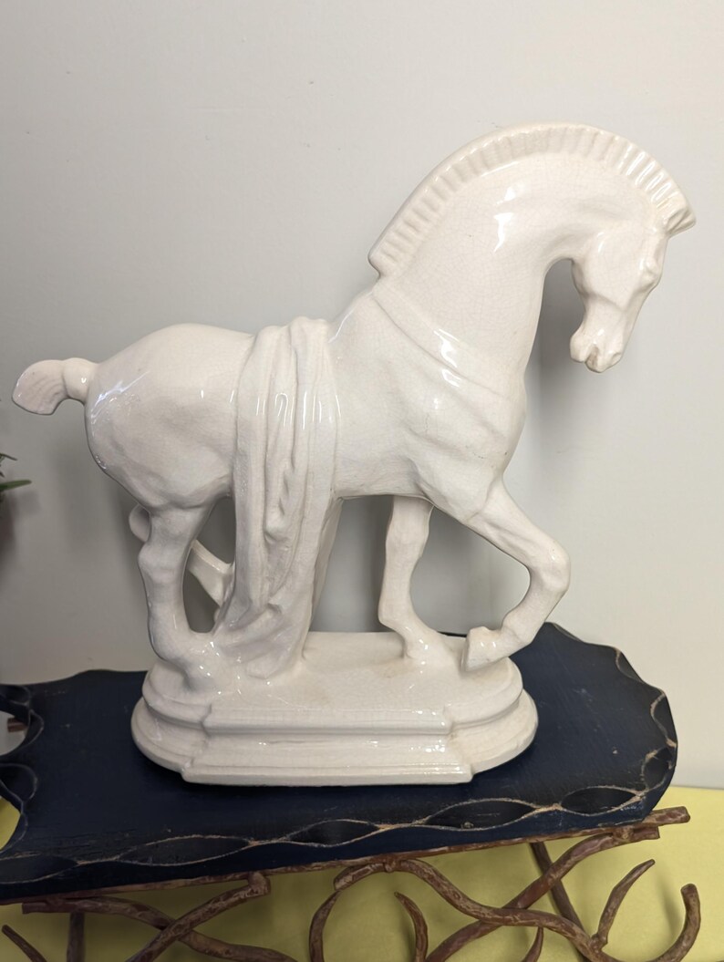 Puede incluir: Figura de caballo de cer&aacute;mica blanca con detalle de tela drapeada. El caballo est&aacute; posado en una postura din&aacute;mica, montado sobre una base rectangular. La figura tiene un acabado brillante y es un art&iacute;culo decorativo.