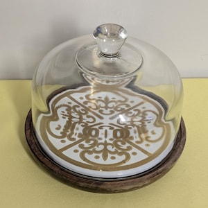 May include: A clear glass cloche with a round knob on top, resting on a white and gold patterned plate. The plate sits on a dark wooden base. The cloche is designed to cover and protect food items.