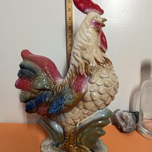 May include: A ceramic rooster figurine with a red comb and wattle, and a multi-colored tail. The rooster is approximately 16 inches tall. The figurine has a textured body and is standing on a base with green and brown accents.