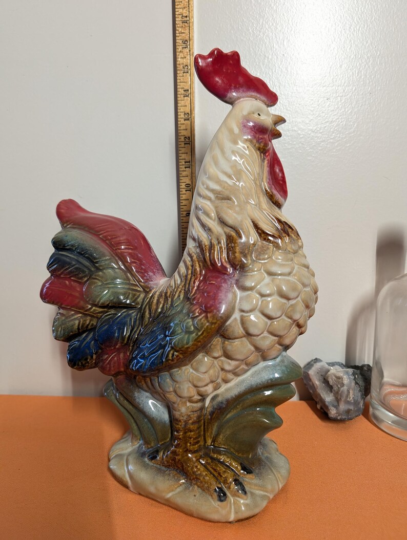 May include: A ceramic rooster figurine with a red comb and wattle, and a multi-colored tail. The rooster is approximately 16 inches tall. The figurine has a textured body and is standing on a base with green and brown accents.