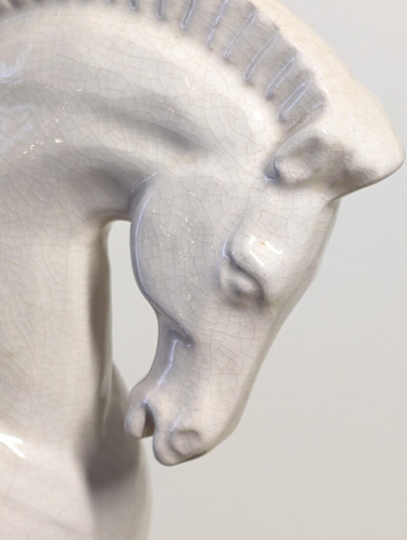 Puede incluir: Escultura de caballo de cer&aacute;mica gris claro. La cabeza del caballo est&aacute; inclinada hacia abajo, mostrando detalles y la superficie texturizada de la cer&aacute;mica. La escultura tiene un acabado craquelado.