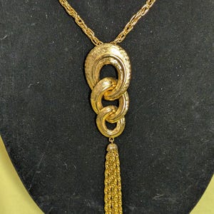 May include: Gold-toned necklace with a textured pendant of interlocking rings and a tassel. The chain is a delicate rope style, and the pendant has a detailed, ornate design. The tassel hangs below the rings, adding movement to the piece.