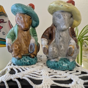 May include: Two ceramic figurines of Peter Rabbit characters wearing hats and holding umbrellas. The figurines are painted in shades of brown, green, yellow, and blue.
