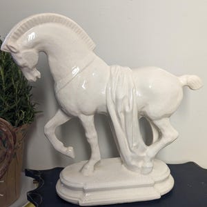 Puede incluir: Una escultura de caballo de cer&aacute;mica blanca con detalle de tela drapeada. El caballo est&aacute; posado con una pata levantada, de pie sobre una base decorativa. Un elemento decorativo para el hogar.
