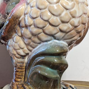 May include: A ceramic rooster figurine with a detailed, textured body in shades of beige and brown. The rooster's feathers are intricately designed, with accents of red, green, and blue. The base and feet are a mix of brown and green.
