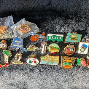 May include: A collection of 18 enamel pins featuring various designs, including flags, landmarks, and logos. The pins are gold-toned and have colorful enamel details. Some of the pins feature text, such as "Alaska the Great Land", "USAFA", "San Pietro Citta del Vaticano", and "Staffs".