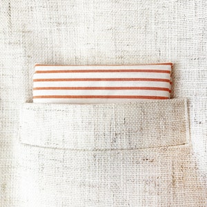 May include: A white linen pocket square with orange stripes. The pocket square is folded and placed in a pocket.