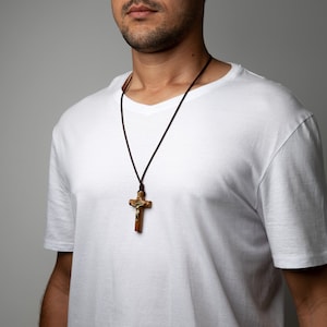 Pode incluir: Um colar de cord&atilde;o de couro marrom com um pingente de crucifixo de madeira. O crucifixo apresenta uma figura de Jesus dourada.