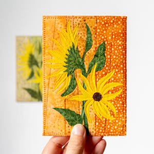 Op de afbeelding: Een handgemaakt stoffen kunstwerk met twee gele zonnebloemen met groene bladeren tegen een oranje achtergrond. Het kunstwerk is gestikt met een decoratieve rand en wordt door een hand vastgehouden. Een tweede, vergelijkbaar stuk staat op de achtergrond.