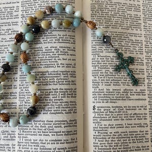 May include: A rosary featuring light blue, tan, and dark brown beads, interspersed with small metallic spacers, resting on an open book with printed text. A dark, textured cross pendant hangs from the end of the beaded strand.