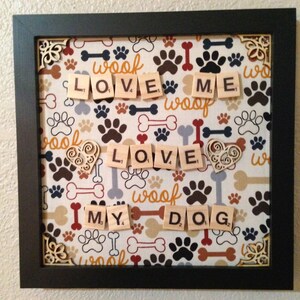 May include: Framed artwork with a dog theme. The background features a pattern of paw prints, bones, and the word "woof." The words "LOVE ME MY DOG" are spelled out with wooden letter tiles. Decorative wooden corner accents.