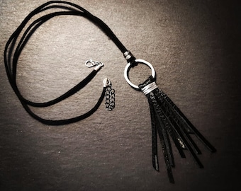 Puede incluir: Un collar de cuero negro con un colgante de anillo plateado y una larga borla de flecos. El collar está sobre una superficie gris oscura.
