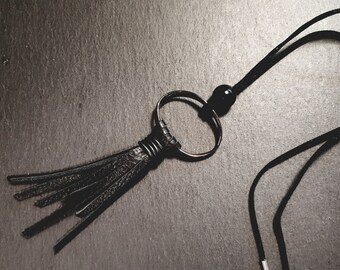 Puede incluir: Collar con borla de cuero negro con un gran anillo de metal y un cordón negro. La borla está envuelta con alambre negro fino. El collar tiene una pequeña cuenta negra y un cierre plateado.