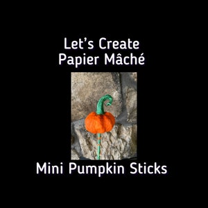 May include: An orange papier-mâché pumpkin with a green stem on a stick. The text "Let's Create Papier Mâché" and "Mini Pumpkin Sticks" are displayed in white text on a black background. The pumpkin is set against a stone wall.