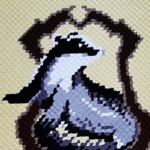 May include: A yellow crocheted blanket featuring a badger design and the words "Hufflepuff" in black. The blanket has a black border and the words "Quizzical Crochet" in the bottom right corner.