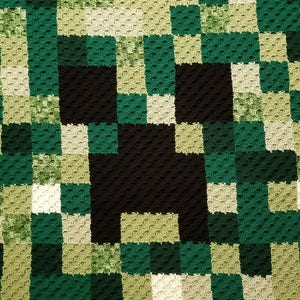 Pode incluir: Uma manta de crochê verde, preta e branca com um design de rosto de Creeper do Minecraft pixelado. A manta tem uma borda de crochê. O texto "Quizzical Crochet" está no canto inferior direito.
