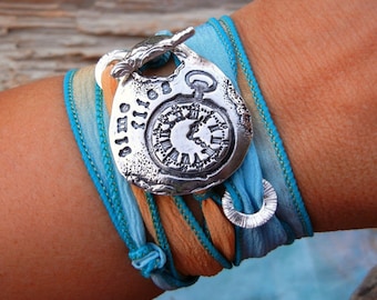 May include: A silver charm bracelet with a clock face charm and the word "time" engraved on it. The bracelet is made of turquoise and orange silk ribbon.