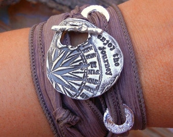 May include: A brown fabric bracelet with a silver charm that says "Enjoy the Journey". The charm has a starburst design and a small loop.