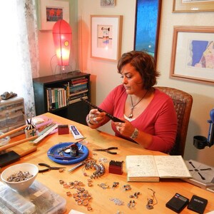 May include: A woman sits at a table with a variety of jewelry making supplies, including beads, wire, and tools. She is working on a piece of jewelry, and there is a notebook open in front of her.