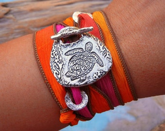 May include: A silver charm bracelet featuring a detailed turtle design, wrapped in orange, pink and brown silk ribbon.