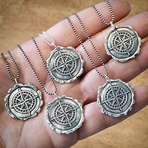 May include: Five silver necklaces with compass pendants. Each pendant features a detailed compass design with a star in the center. The necklaces are on a hand with a light brown background.