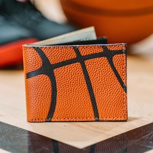 May include: A bi-fold wallet with a basketball design. The wallet is orange with black lines mimicking a basketball's pattern. The wallet is made of textured material and appears to be new.