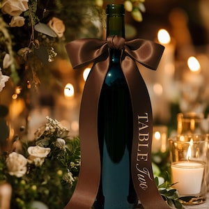 May include: A dark green wine bottle decorated with a brown satin ribbon tied in a bow. The ribbon has the words "Table Two" printed on it. The background includes candles and floral decorations, creating a warm ambiance.