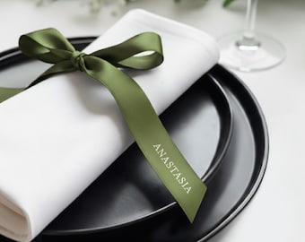 May include: A white cloth napkin folded and tied with an olive green satin ribbon. The ribbon features the name ANASTASIA printed in white capital letters. The napkin rests on a matte black plate, which is set upon a larger black plate.