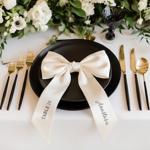 May include: A formal table setting featuring a black plate, gold and black cutlery, and a cream bow with the name "Anastasia" and "Table 10". White flowers and foliage are arranged in the background.