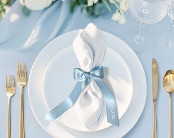 May include: A white cloth napkin folded and secured with a light blue satin ribbon tied in a bow. The ribbon features the name Taylor printed in white lettering. The napkin rests on a white plate, flanked by gold-toned forks, a knife, and a spoon.