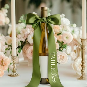 May include: A wine bottle adorned with a green satin ribbon bow and a long streamer with the words "Table Two" in white. The bottle is surrounded by candles and a floral arrangement with pink and white roses, creating an elegant table setting.