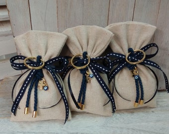 Op de afbeelding: Drie kleine beige stoffen zakjes met een blauw-wit lint eromheen gebonden. Elk zakje heeft een gouden ring met een bedel eraan. De bedels omvatten een blauw boze oog, een gouden kruis en een blauw en wit boze oog.