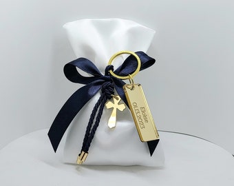 Op de afbeelding: Een witte satijnen tas met een donkerblauw lint en een gouden sleutelhanger met een kruisbedel en een rechthoekig label met de tekst "Eloise 01.13.2023".