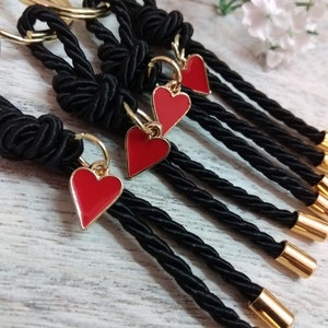 May include: Five black rope keychains with gold rings and red heart charms. The keychains are tied with a knot at the top and have gold beads at the bottom.