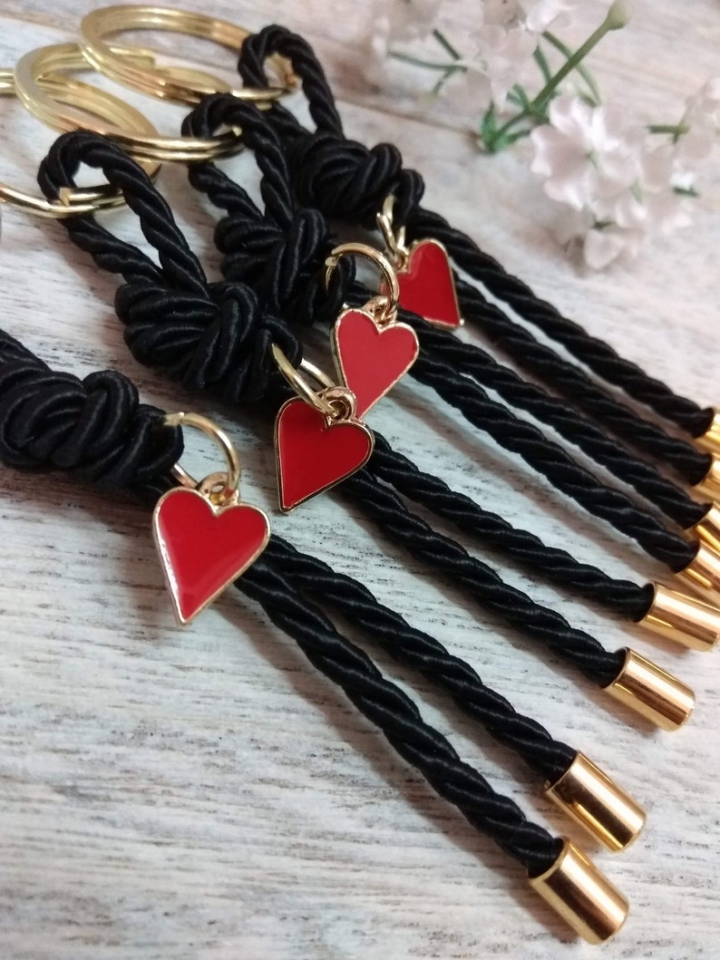 May include: Five black rope keychains with gold rings and red heart charms. The keychains are tied with a knot at the top and have gold beads at the bottom.