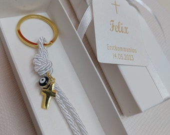 Op de afbeelding: Een witte geschenkdoos met een gouden sleutelhanger erin. De sleutelhanger heeft een wit touw met een gouden kruis en een blauwe kwade oog-bedel. De doos is versierd met een wit lint en een label met de tekst "Felix Eerste Communie 14.05.2023".