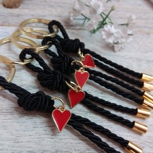 May include: Five black rope keychains with gold rings and red heart charms. The keychains are made of braided rope and have gold tips.