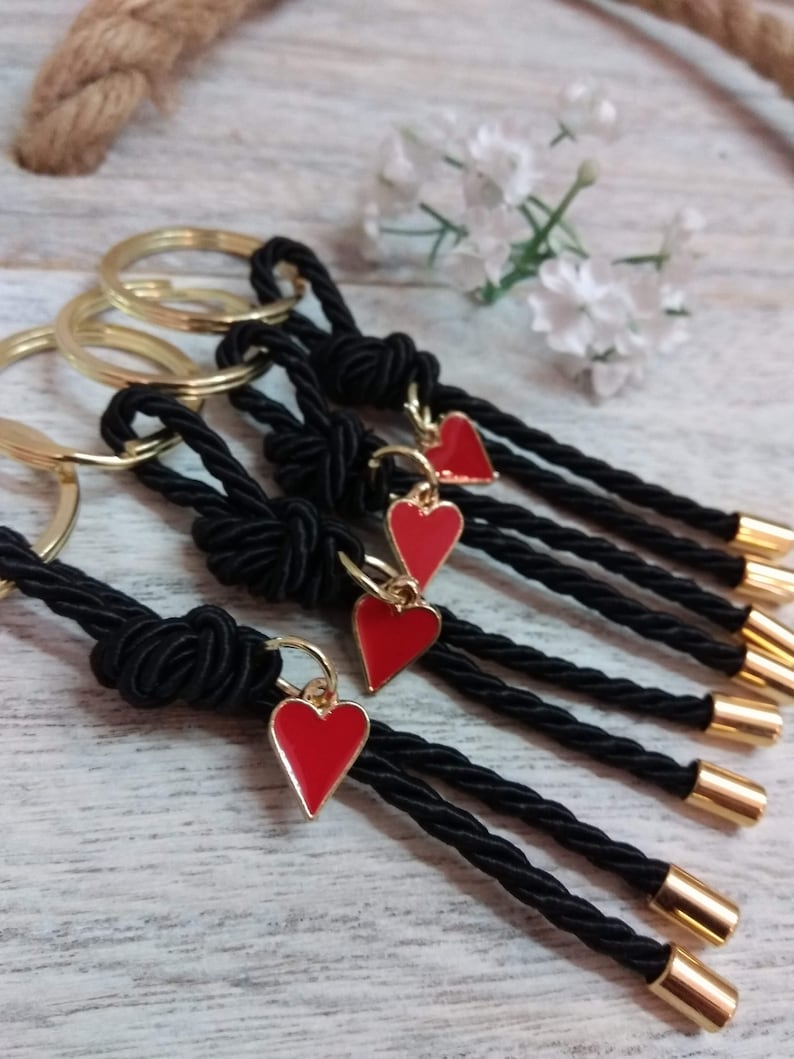 May include: Five black rope keychains with gold rings and red heart charms. The keychains are made of braided rope and have gold tips.