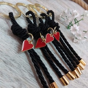 May include: Five black keychains with gold rings and red heart charms. Each keychain has a black tassel with a gold tip.