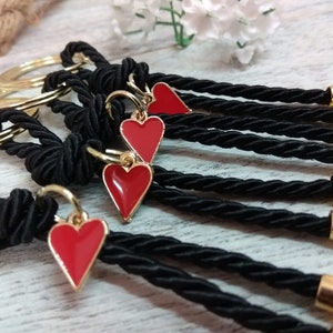 May include: A set of five black keychains with gold rings and red heart charms. Each keychain has a black braided cord with a gold bead at the end.