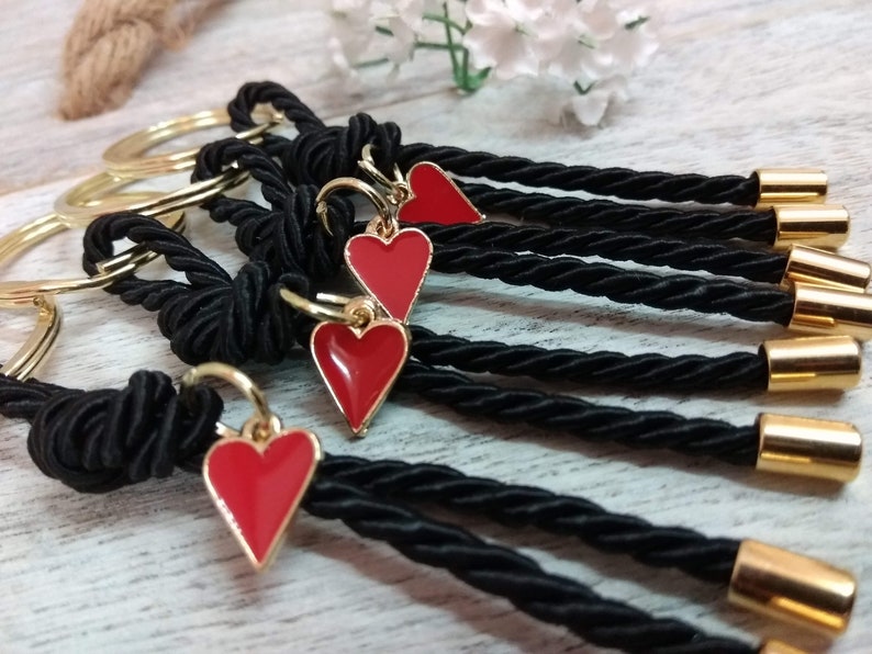 May include: A set of five black keychains with gold rings and red heart charms. Each keychain has a black braided cord with a gold bead at the end.